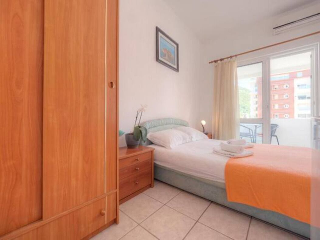 Ein Schlafzimmer mit einem Bett, einem Nachttisch, einem Kleiderschrank, einem Fenster mit Vorhängen und Blick auf Gebäude.