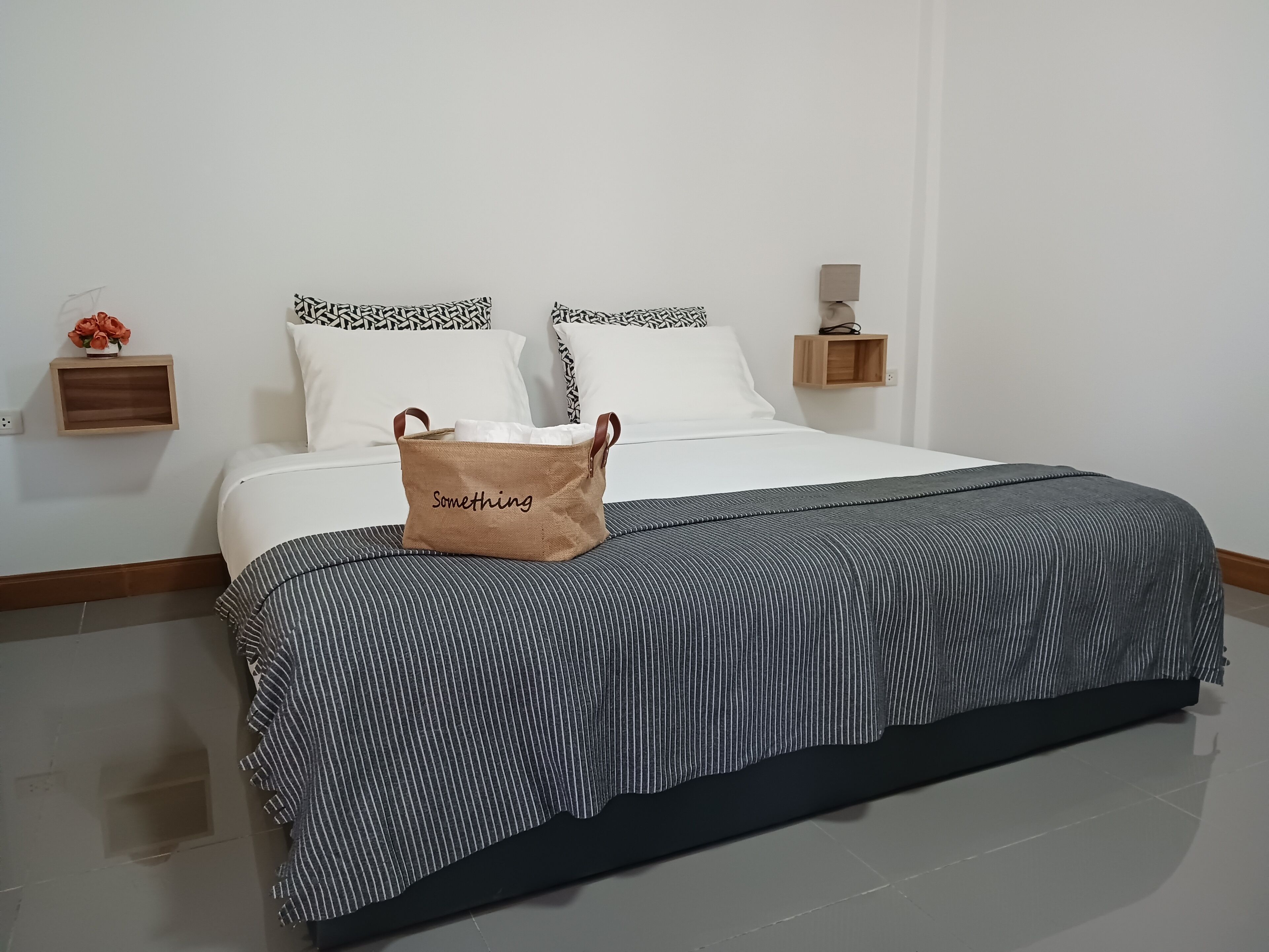 A neatly made bed with a striped bedspread, a brown bag labeled "Something" on the bed, and a small wooden shelf with a vase of flowers.