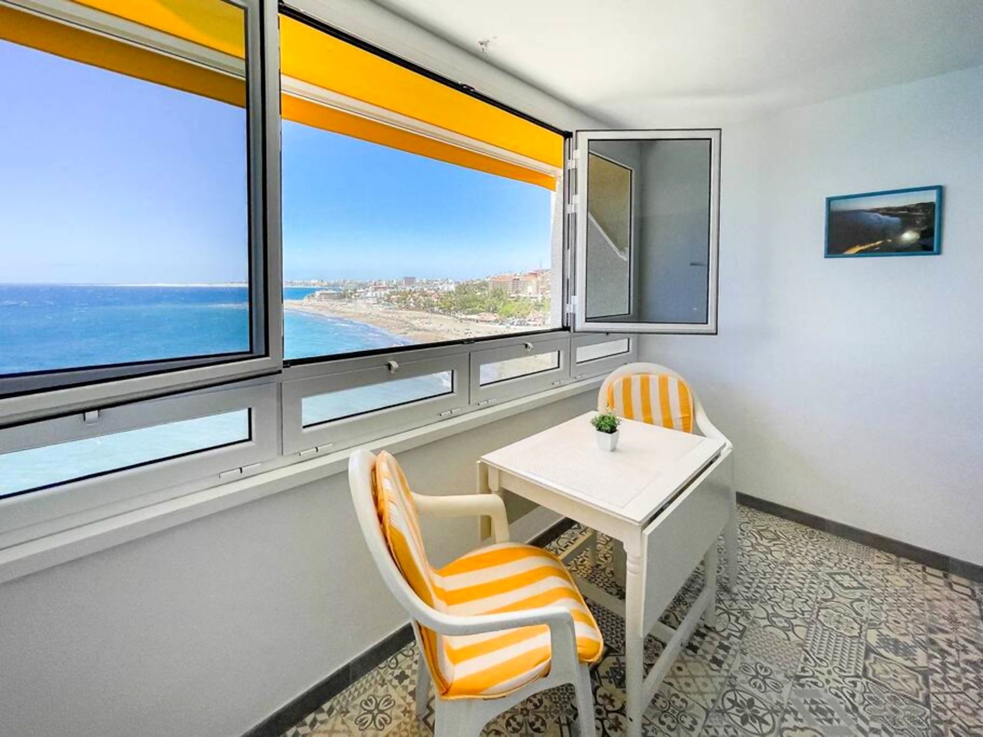 A room with a view of the ocean, a small table, two striped chairs, and a framed picture on the wall.