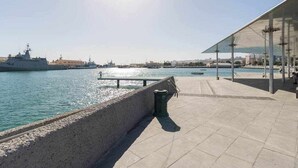 Eine Uferpromenade mit einem großen Schiff im Hintergrund, blauem Himmel und einem modernen Gebäude mit Glasfassade.