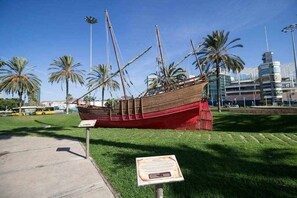 A historical ship model displayed outdoors with informational plaques, surrounded by palm trees and urban buildings.