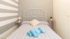 Ein ordentlich bezogenes Bett mit gemusterten Bettwäsche und gerollten Handtüchern, ein Nachttisch mit Lampe und ein kleiner Blumentopf.