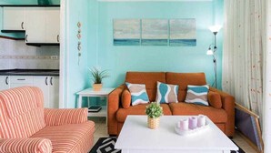 Ein Wohnzimmer mit einem gestreiften Sofa, einem orangefarbenen Sofa mit gemusterten Kissen, einem weißen Couchtisch und einem Meeresblick-Gemälde an der Wand.