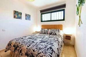 A bedroom with a bed, two wall-mounted lamps, a window with blinds, and two wall-mounted artworks.