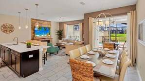 Una cocina moderna con una isla central, comedor con una mesa grande y sillas, y sala de estar con sofá y televisor.