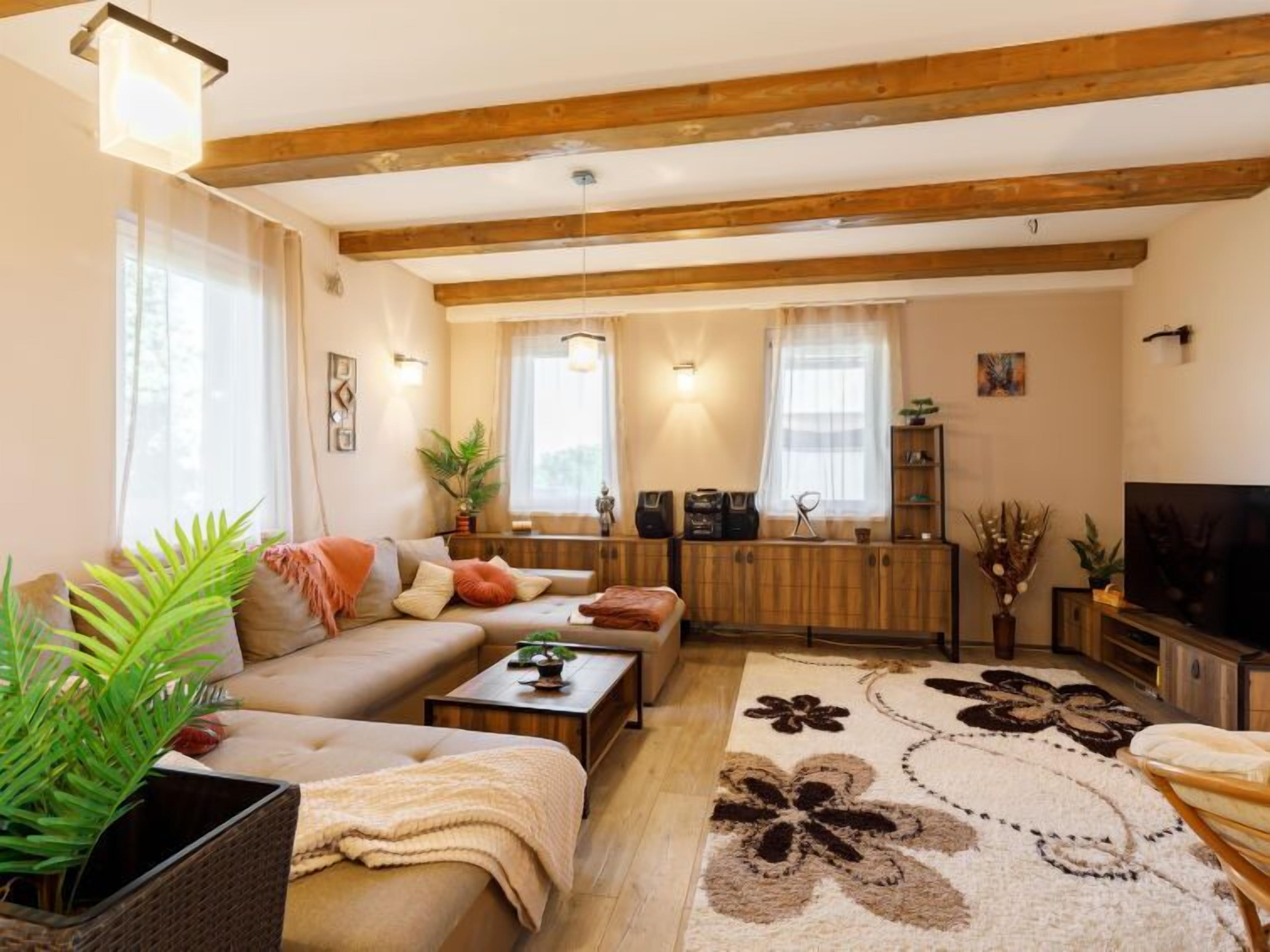 A cozy living room with a sofa, coffee table, and a TV. There are windows with curtains, a fireplace, and a wooden ceiling.
