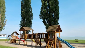 A wooden playground with slides and climbing structures, set on sandy ground with grass in the foreground, and a clear sky in the background.