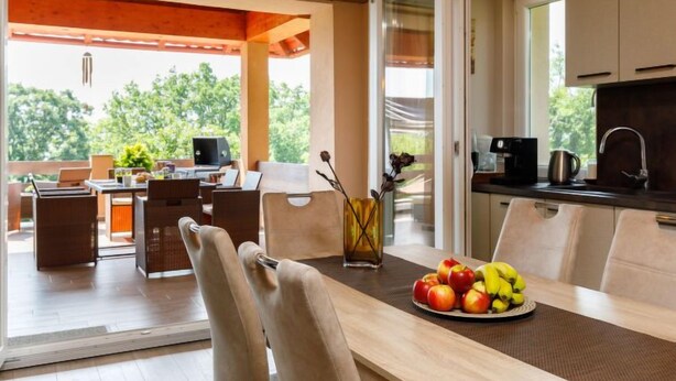 A modern kitchen with a dining area, a view of greenery outside, and a table with fruit.