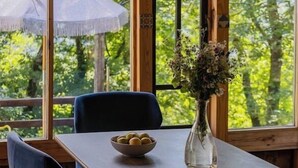 A dining area with a table, chairs, a vase with flowers, and a view of trees through large windows. - Wine Space (Ambrolauri)