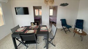Un comedor con una mesa dispuesta para cuatro, sillas, una lámpara colgante, un televisor en la pared y una cama visible a través de una puerta abierta.