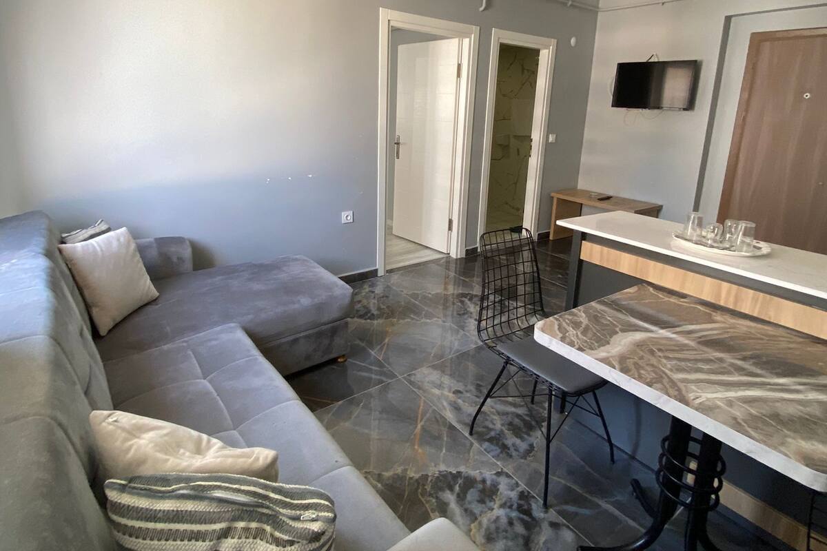 Ein modernes Wohnzimmer mit einem grauen Ecksofa, einem Essbereich mit Marmorfliesen und einem an der Wand montierten Fernseher.