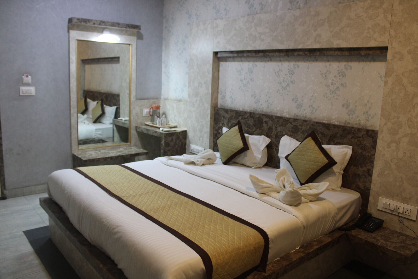 A hotel room with a large bed, a bedside table with a phone, a mirror, and a wall with a floral pattern.