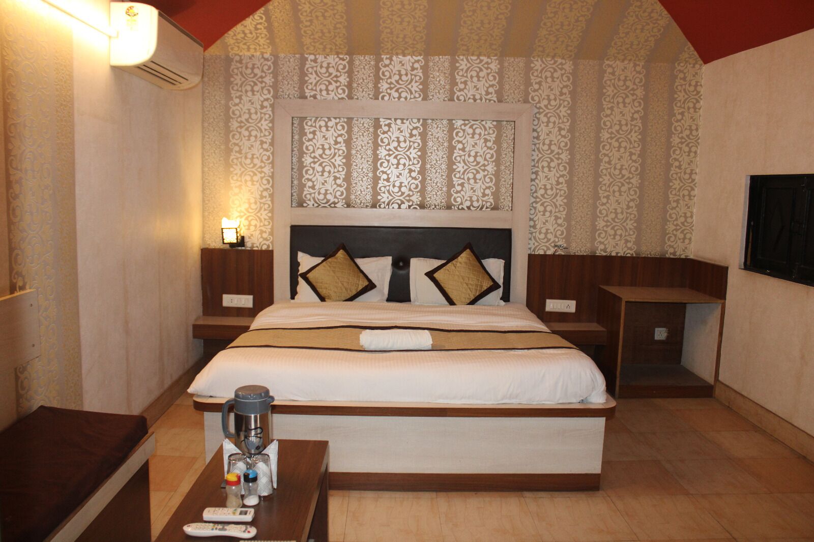 A hotel room with a large bed, two bedside tables, a nightstand, a wall-mounted air conditioner, and a small table with a remote control and a glass.