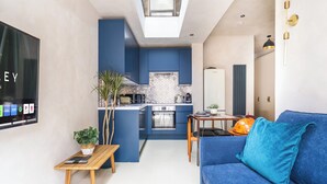 Un salon moderne avec un canapé bleu, une petite table en bois, un téléviseur fixé au mur et une cuisine en arrière-plan.