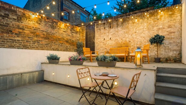 Eine Terrasse im Freien mit Backsteinmauer, Lichterketten, Blumentöpfen und einem Tisch mit Stühlen.
