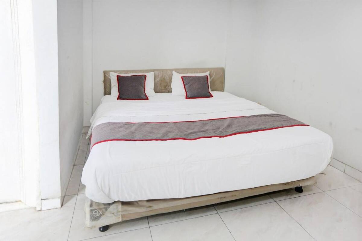 Una cama individual con colchón blanco y detalles rojos, dos almohadas con ribetes rojos y estructura de madera.