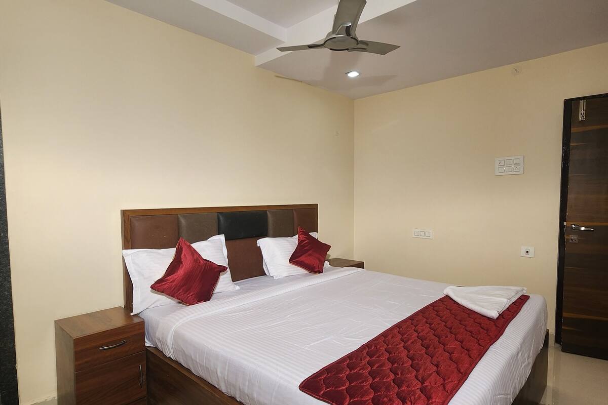 Un lit bien fait, avec une literie blanche et rouge, une tête de lit en bois, une table de chevet, un ventilateur de plafond et une porte.