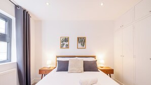 Een net opgemaakt bed met een witte dekbedovertrek, twee kussens en een gele plaid. Twee nachtkastjes met lampen, een raam met gordijnen, en twee ingelijste foto's aan de muur.