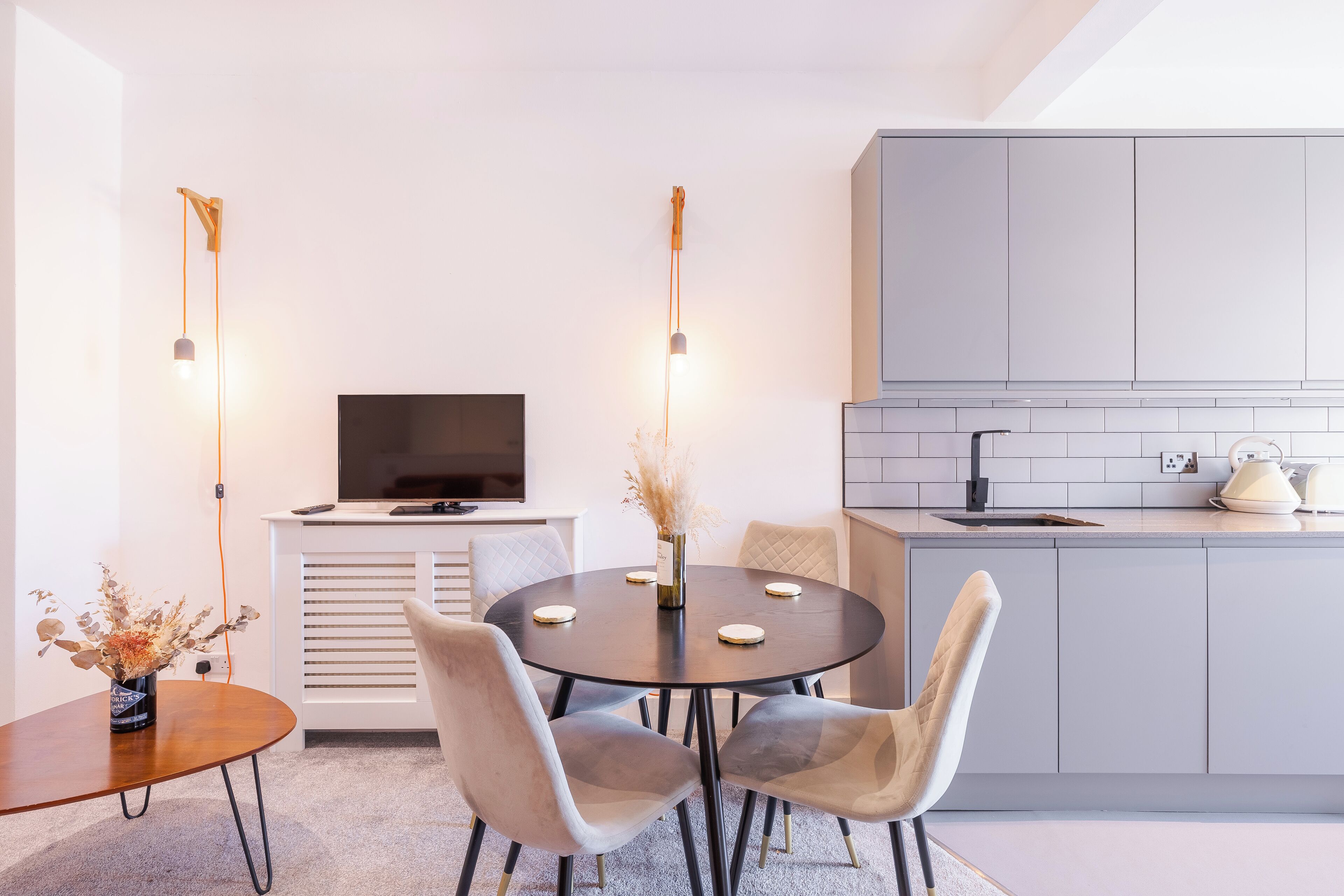 A modern kitchen with grey cabinets, a black countertop, and a built-in oven. There is a dining area with a round table and four chairs, and a TV on a white cabinet.