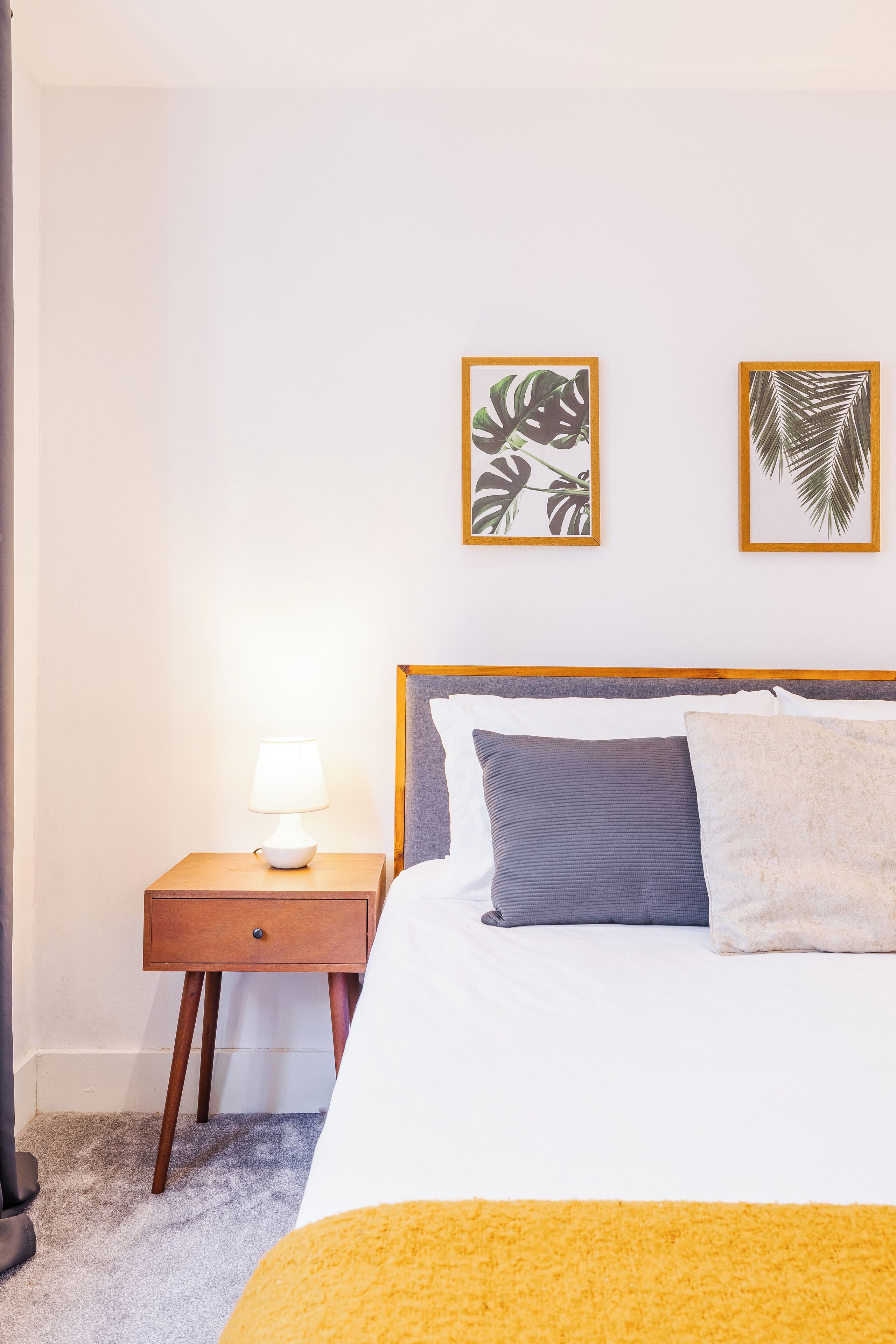 A neatly made bed with a wooden headboard, a bedside table with a lamp, and two framed leaf artworks on the wall.