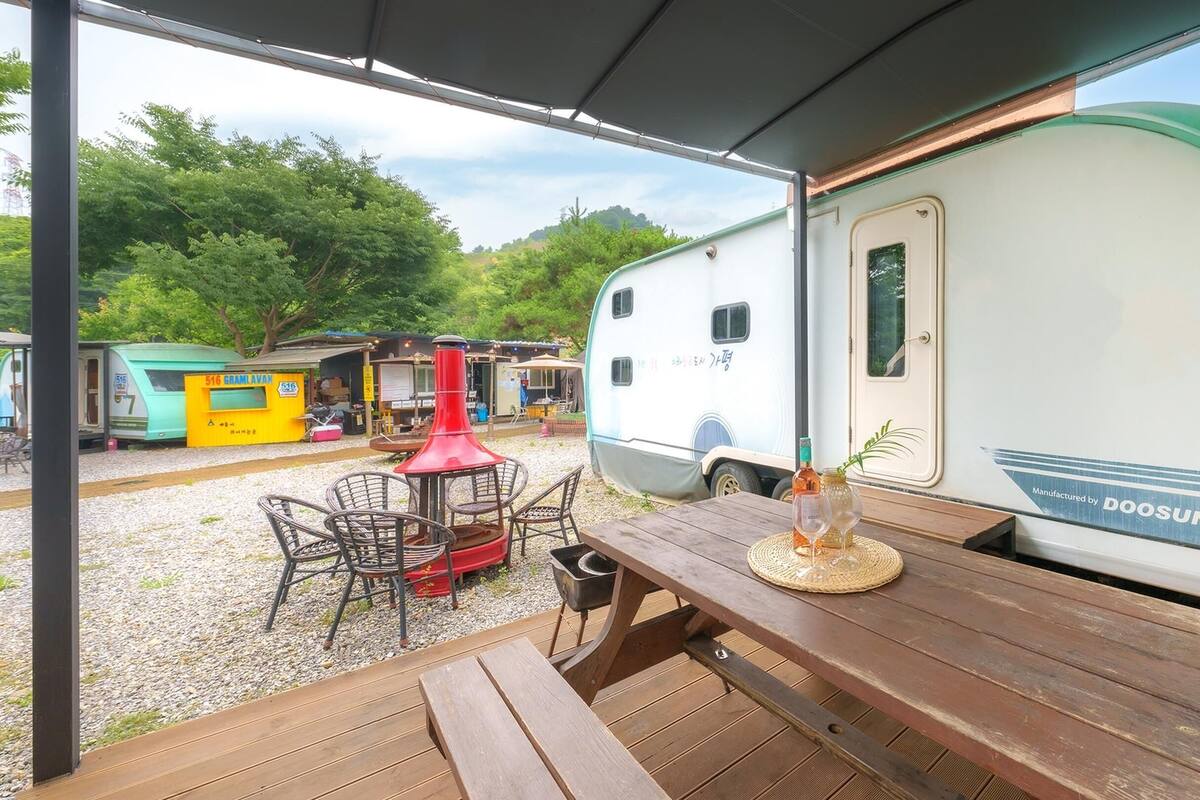 Un espacio al aire libre cubierto con una mesa de picnic de madera, una parrilla de barbacoa roja y una caravana estacionada cerca.