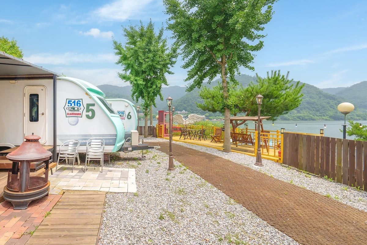 Un camping con una caravana, mobiliario exterior y vistas a un lago y montañas.