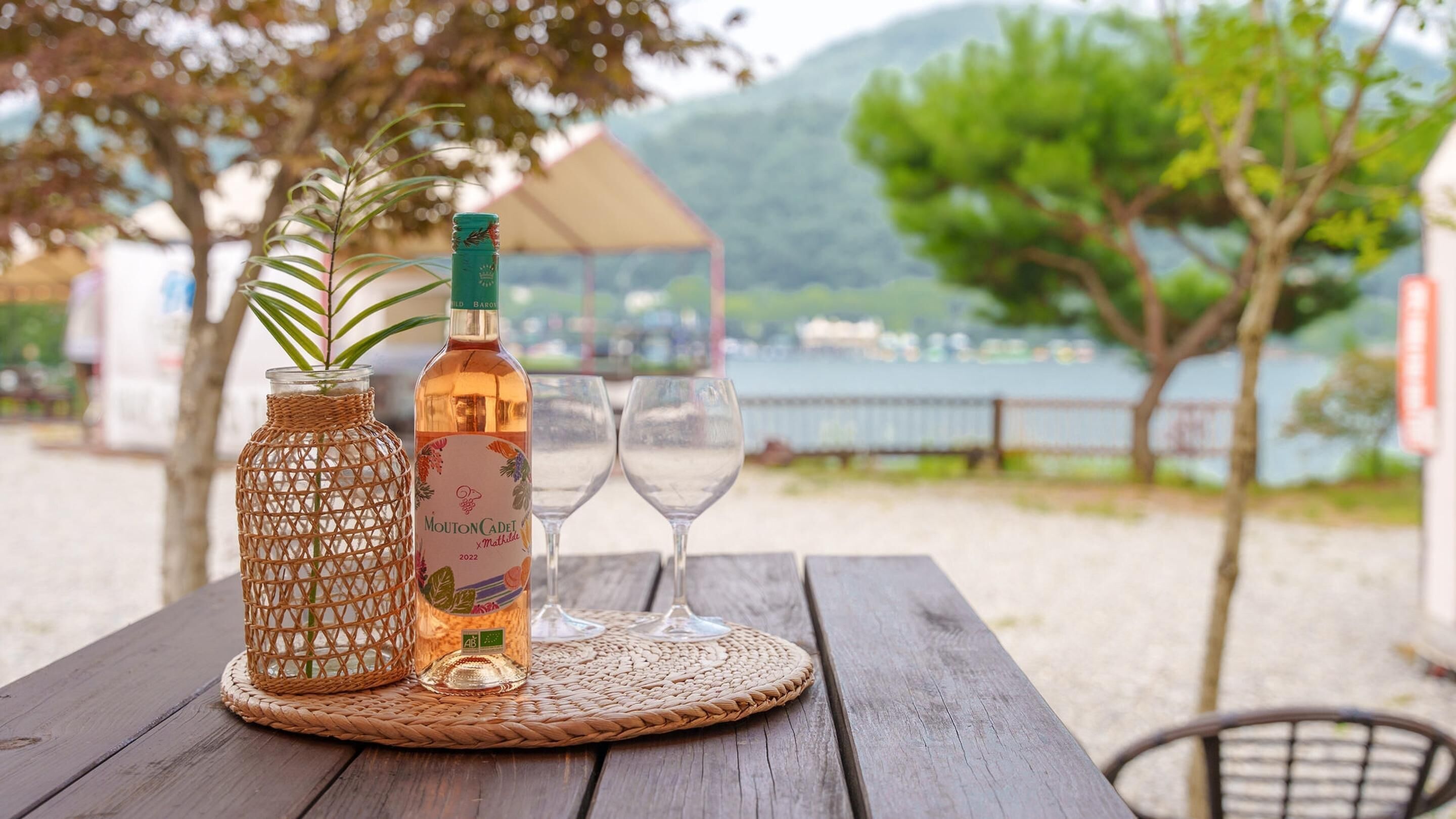 A wooden table with a woven mat, a bottle of Mouton Cadet wine, and two wine glasses.