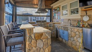 Cocina de piedra y madera con parrilla, asientos de bar y vistas amplias a través de grandes ventanales.