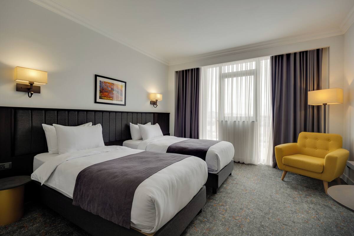 A hotel room with two beds, a chair, a lamp, and a painting on the wall.