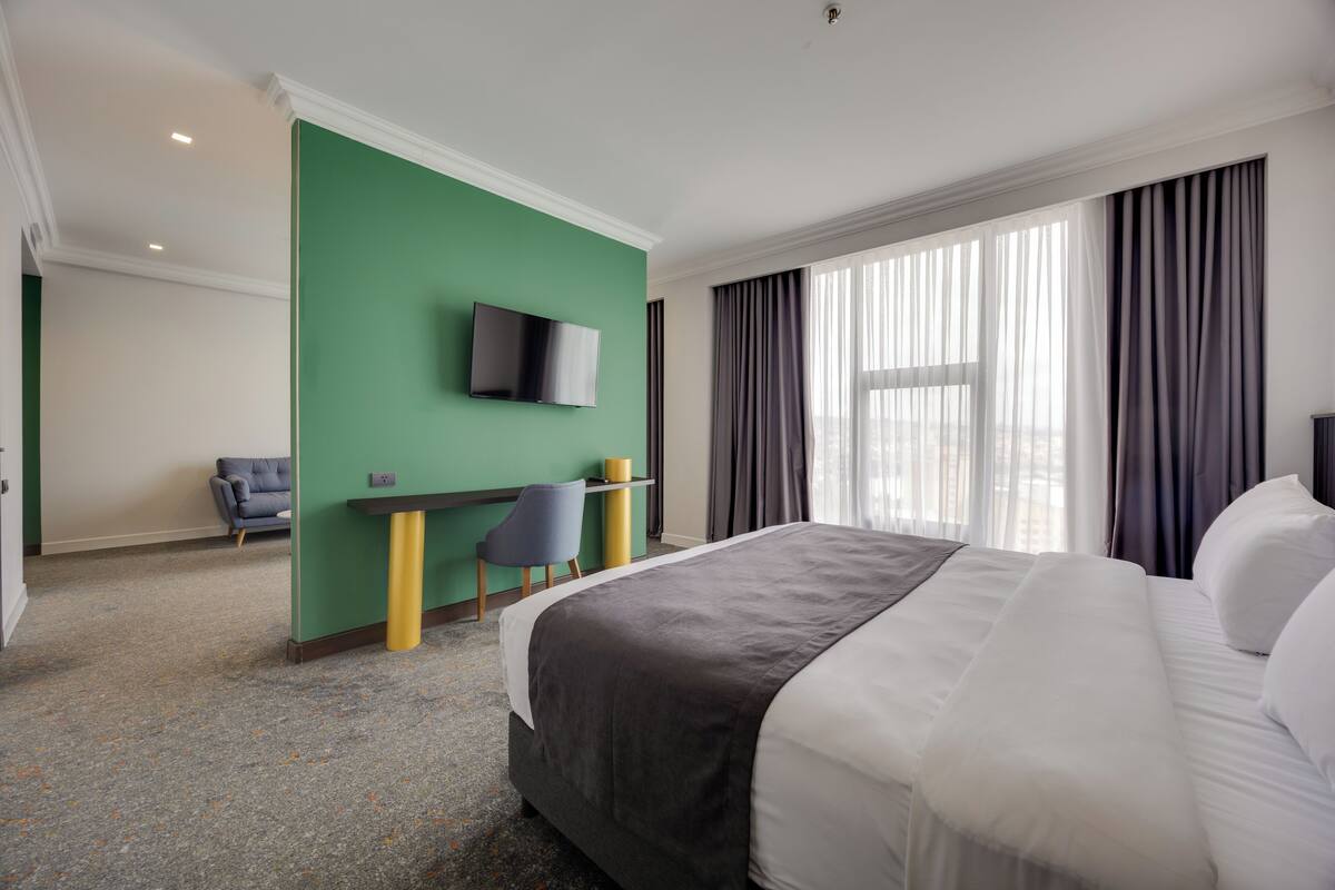 A hotel room with a bed, a desk, a chair, a television, and large windows with curtains.