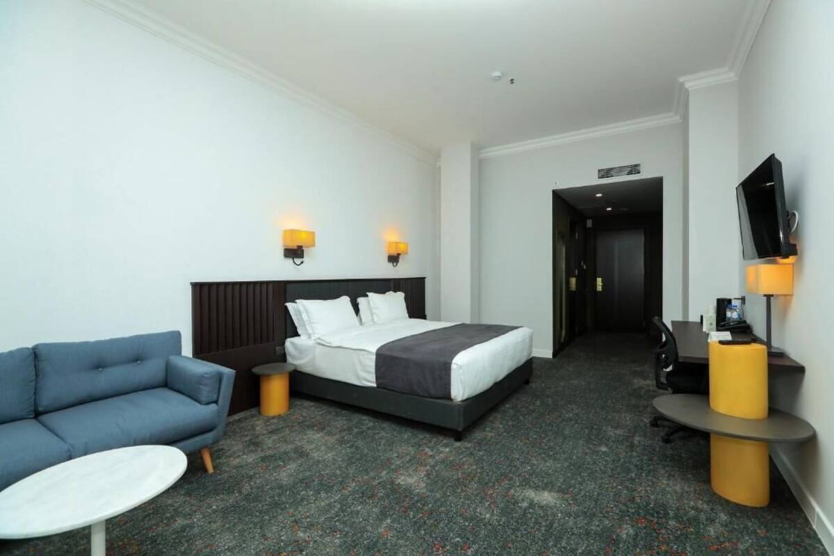 A hotel room with a bed, a sofa, a small table, a TV, and a desk with a lamp.