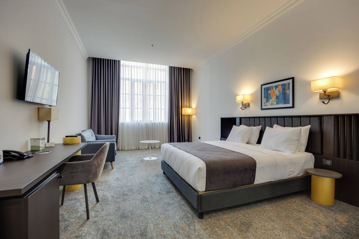 A hotel room with a bed, desk, chair, TV, and artwork on the wall.
