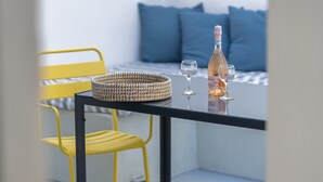 Una mesa con una cesta tejida, una botella y dos vasos en un balcón, con una cama y almohadones azules al fondo.