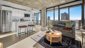 Eine moderne Küche mit einer zentralen Kochinsel, einem Essbereich mit rundem Tisch und einem Wohnbereich mit Sofa und großen Fenstern.