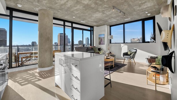 Ein modernes Interieur mit großen Fenstern, einer zentralen Insel mit Schubladen, einer Sitzecke mit Stuhl und Tisch sowie Blick auf die Stadt.