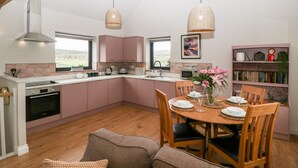 A modern kitchen with pink cabinets, a dining area with wooden chairs, and a bookshelf. - Sevenwoods View (Longhope)