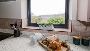 A kitchen with a window, a countertop, a teapot, cups, a loaf of bread, and a cutting board with grapes and strawberries. - Sevenwoods View (Longhope)