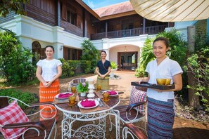 Une cour intérieure avec une table à manger dressée pour un repas, entourée d’une végétation luxuriante et bordée d’un bâtiment doté d’un balcon.
