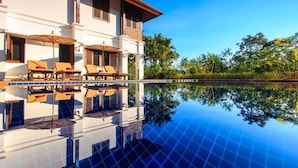 A hotel with a pool reflecting the building and surrounding trees, lounge chairs, and a clear blue sky.