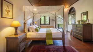 A bedroom with a four-poster bed, wooden flooring, a nightstand with a lamp, a dresser, and a large window with a view of greenery.