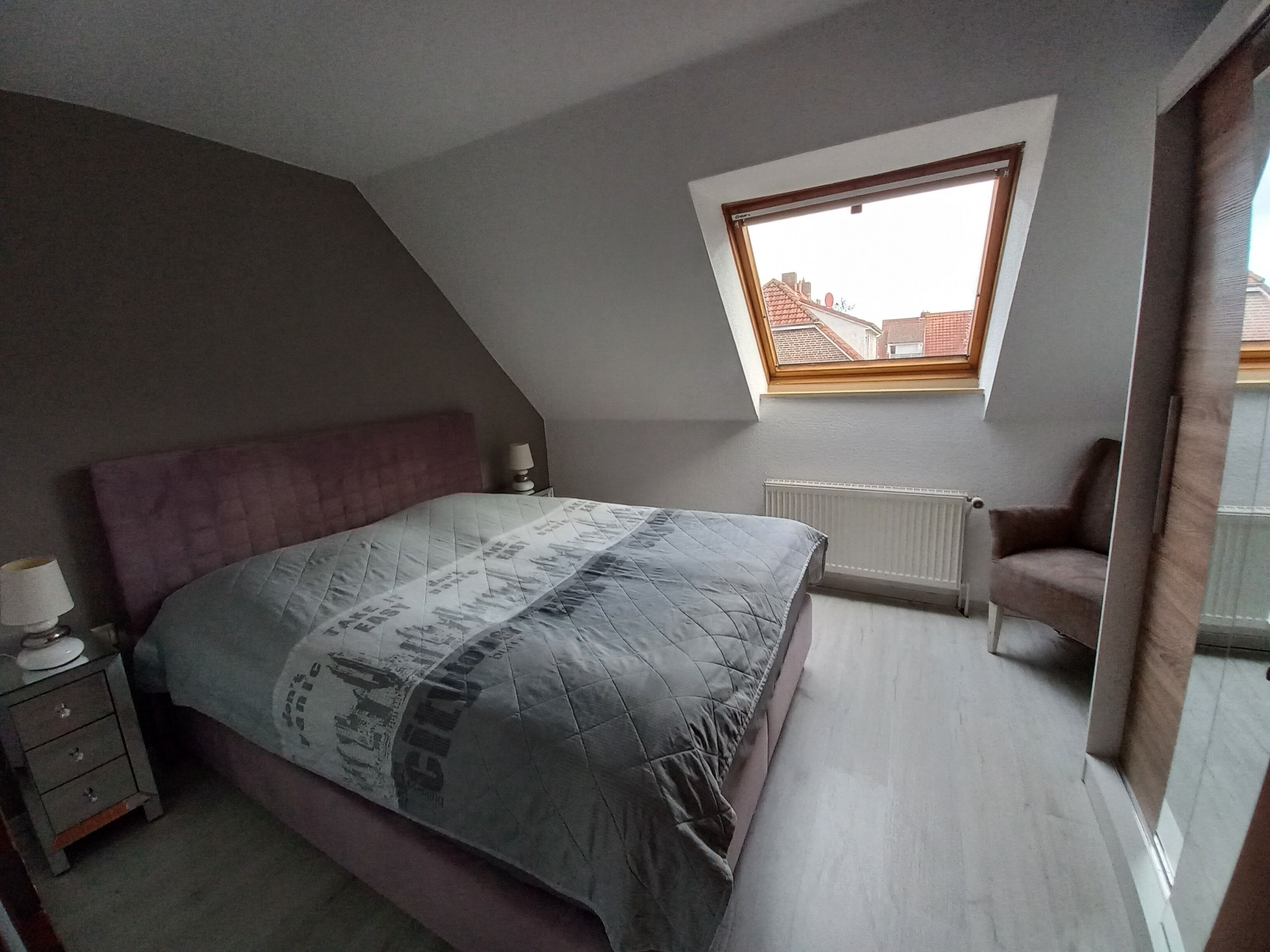 Een slaapkamer met een schuine plafond, een bed met een beddegoed met een patroon, een nachtkastje met een lamp en een raam met uitzicht op gebouwen.
