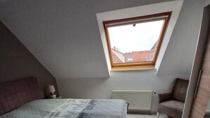 A bedroom with a sloped ceiling, a skylight, a bed with a patterned duvet, a radiator, and a chair.