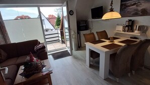 A modern living room with a sofa, coffee table, and dining area. There is a TV mounted on the wall, a ceiling fan, and a painting above the dining table.