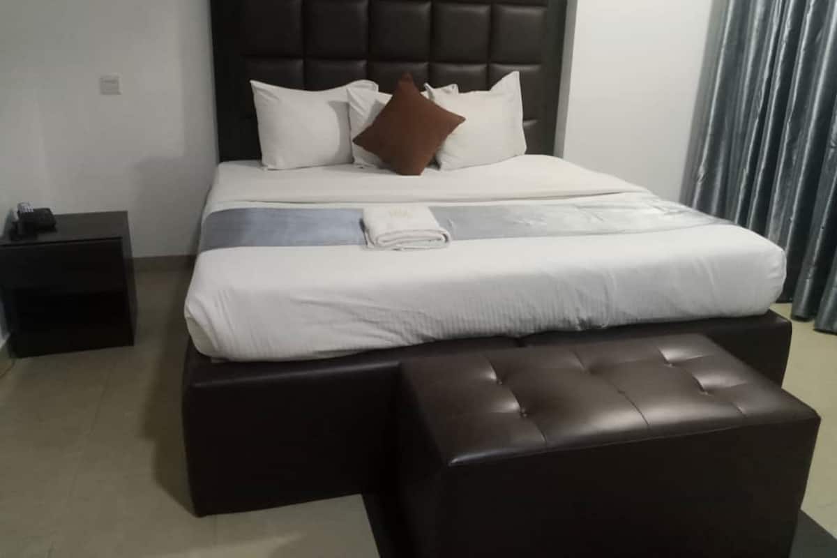 A neatly made bed with white and brown pillows, a grey blanket, and a brown ottoman in a hotel room with a dark headboard and a small black nightstand.