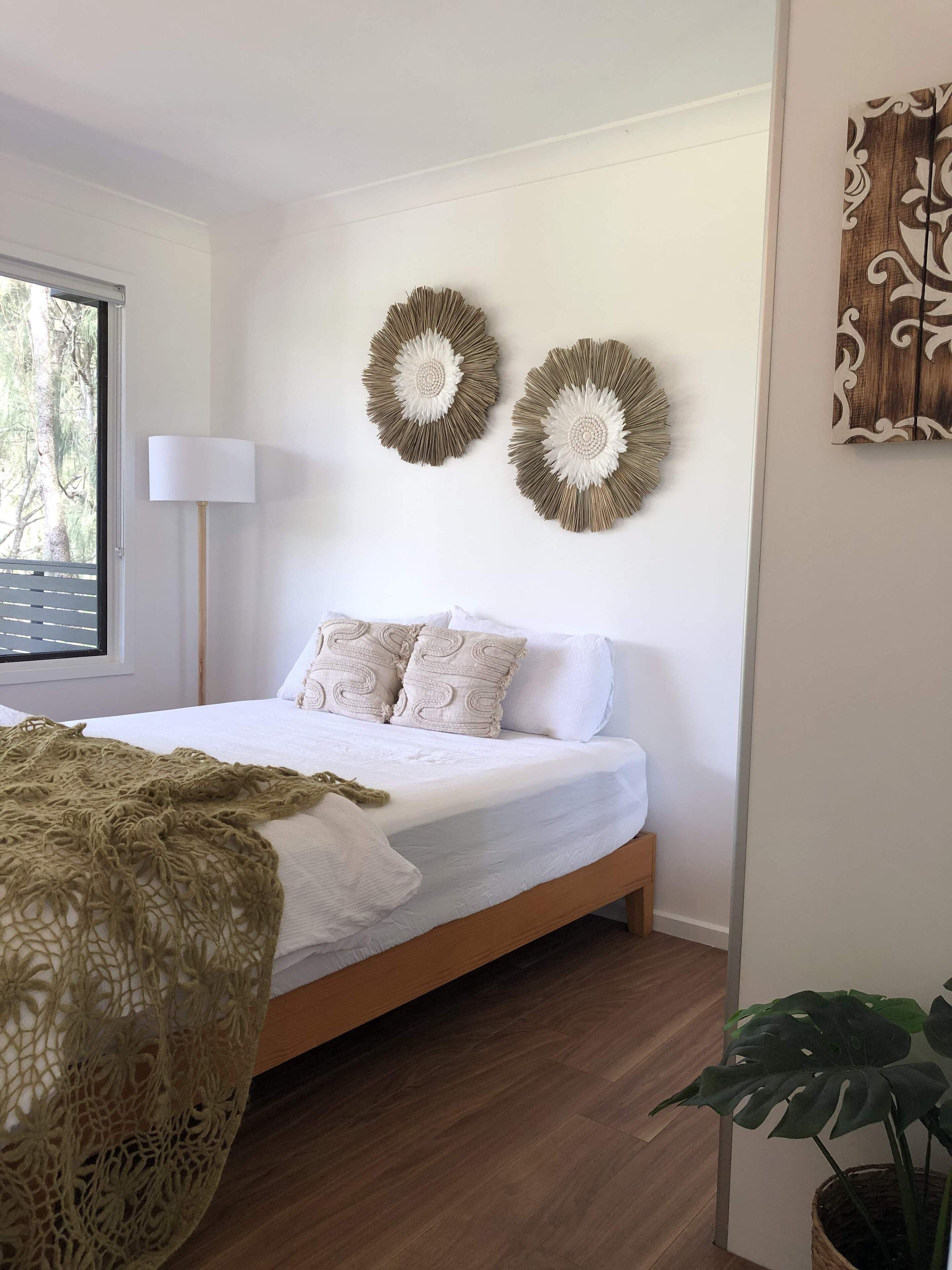 Une chambre à coucher comprenant un lit, deux éléments de décoration murale, une lampe sur pied, une couverture tissée et une plante en pot.