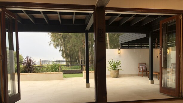 Un patio abierto con vistas a un jardín, una mesa de madera con un bol, y una planta en maceta.