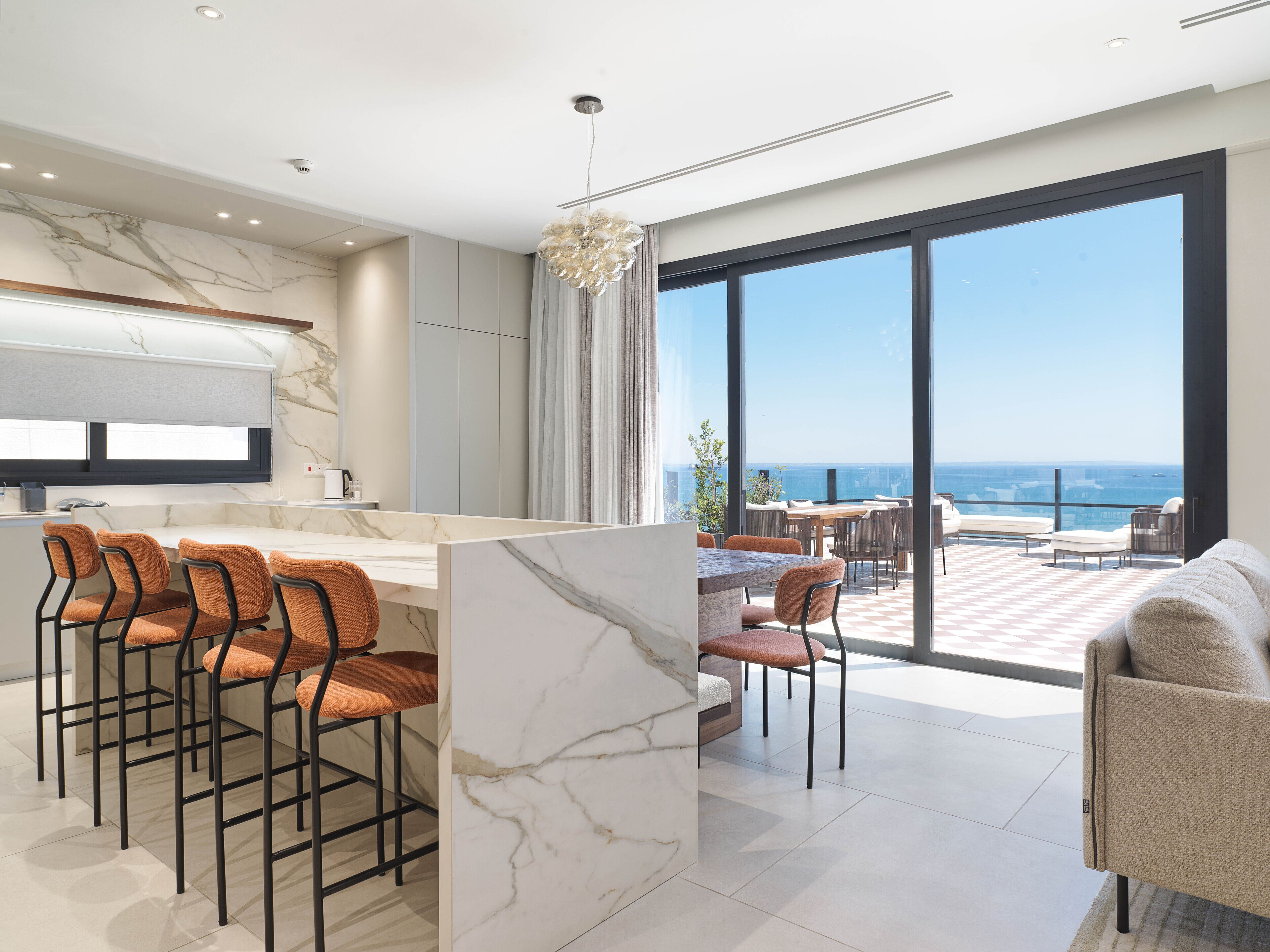 A modern kitchen with marble countertops, a bar area with stools, and a large window overlooking the sea.