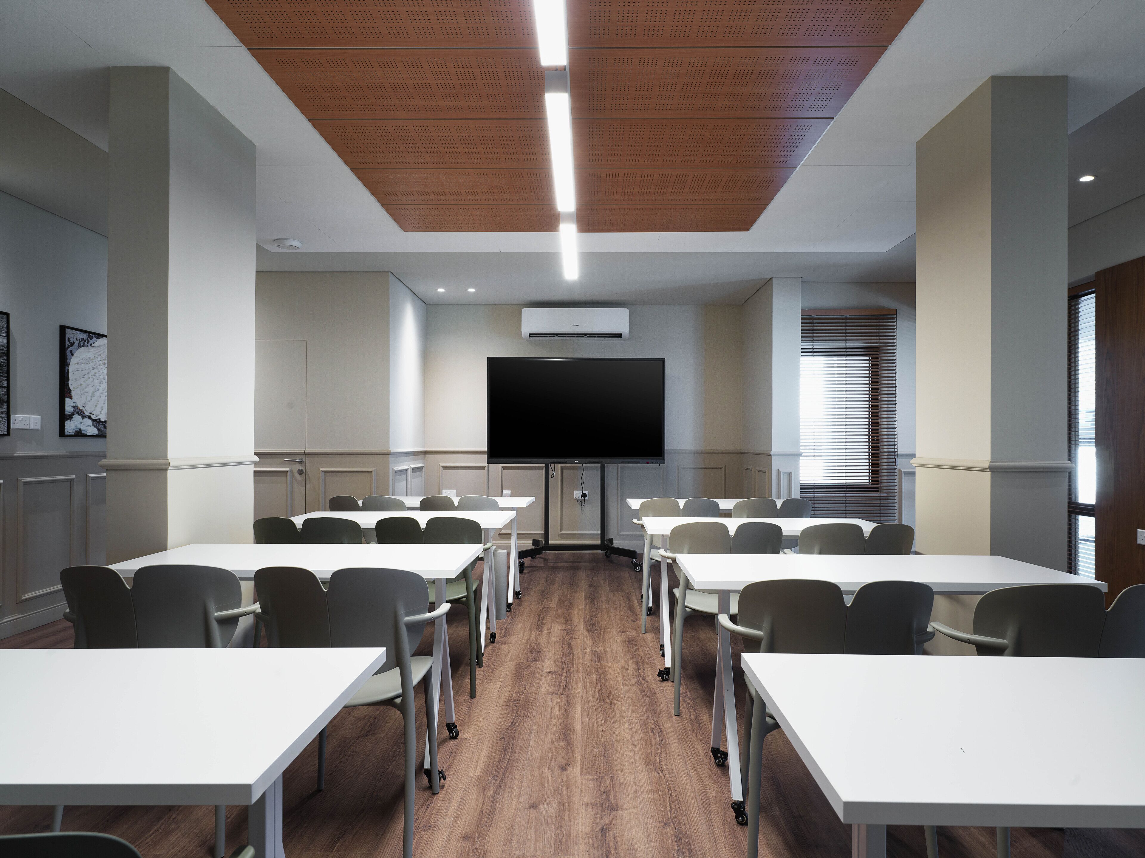 Une salle de conférence avec des tables et des chaises blanches, un grand téléviseur à écran plat et un climatiseur fixé au mur.