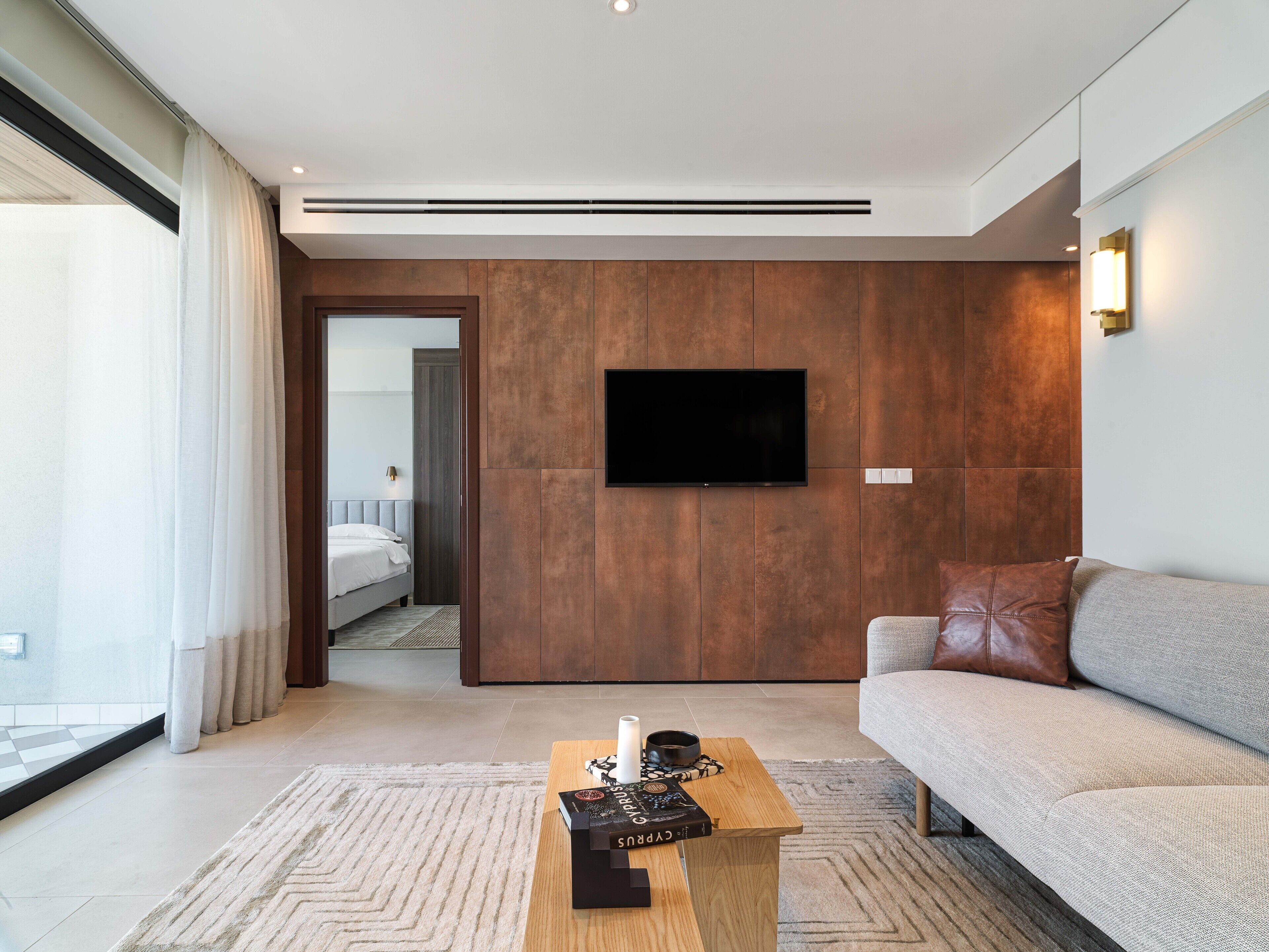 Una habitación de hotel moderna con un televisor de pantalla plana grande montado en una pared de madera, un sofá, una mesa de centro de madera con un libro, y vista de la habitación contigua a través de una puerta de cristal.