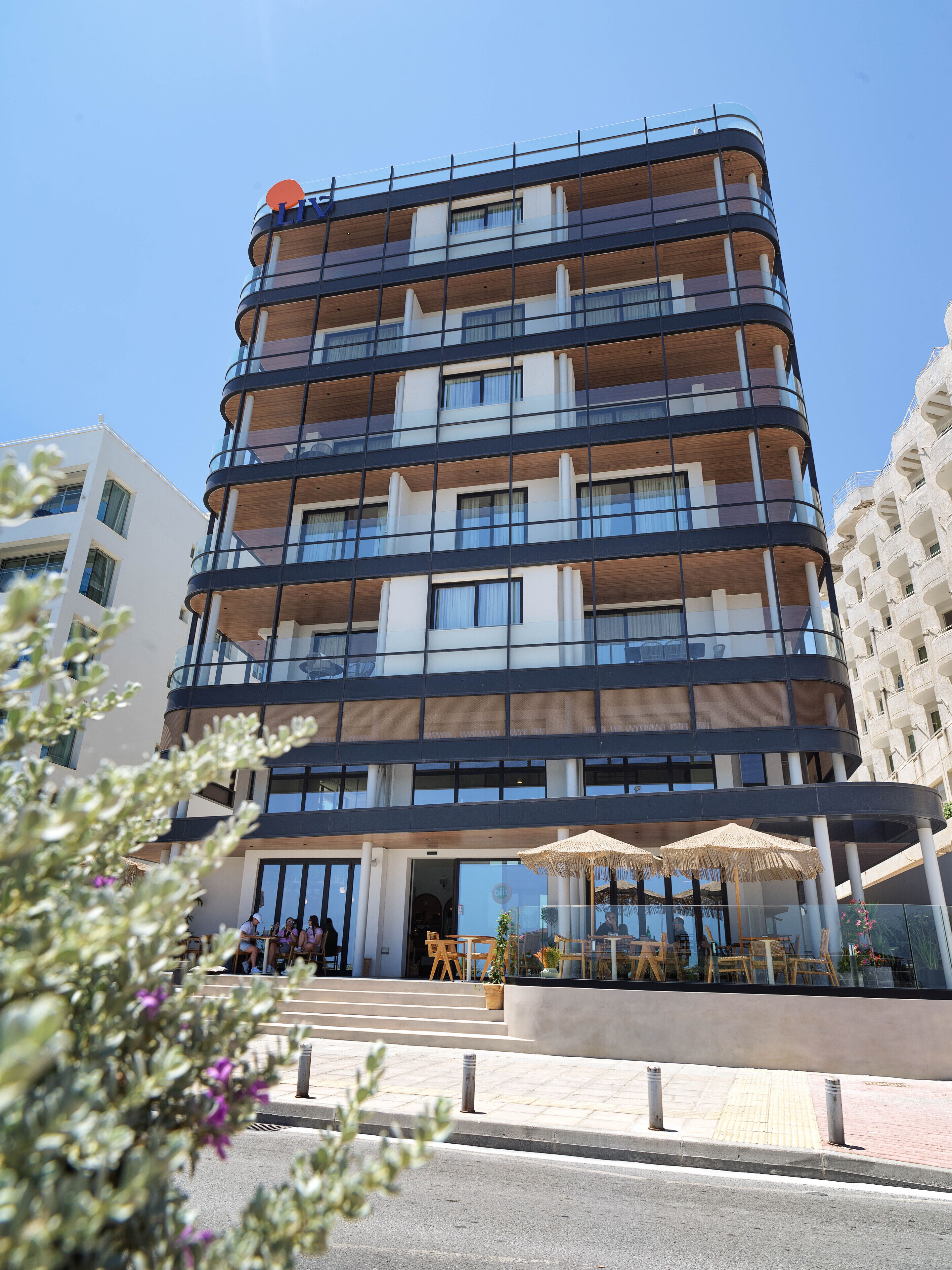 A multi-story hotel with a modern design, featuring balconies, large windows, and a ground-level outdoor seating area with umbrellas.